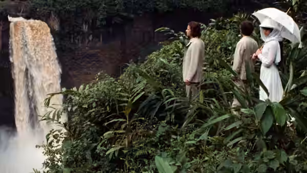 Three travelers stand among dense foliage beside a waterfall, one holding a parasol