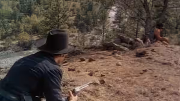 A black-hatted man crouches with a revolver, watching the rocky ground ahead