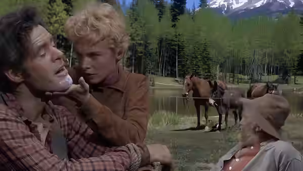 A blonde woman holds a dark-haired man as two horses stand beside a lakeside camp