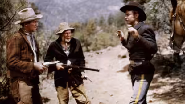 A gunman with a rifle faces a black-clad man raising his hands in a clearing