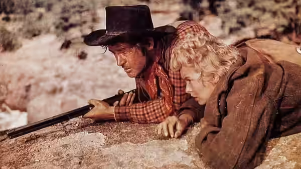 A man and blonde woman crouch behind a rocky ledge, aiming a rifle