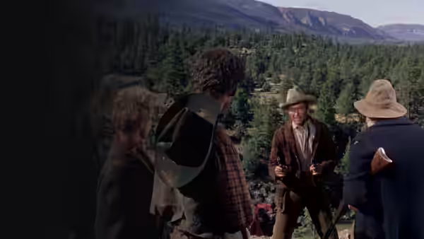 Four men gather near a mountain camp, with horses and forested slopes behind them