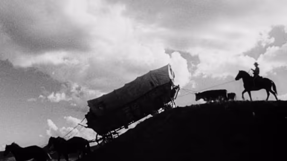 A covered wagon climbs a ridge as a rider on horseback watches from above