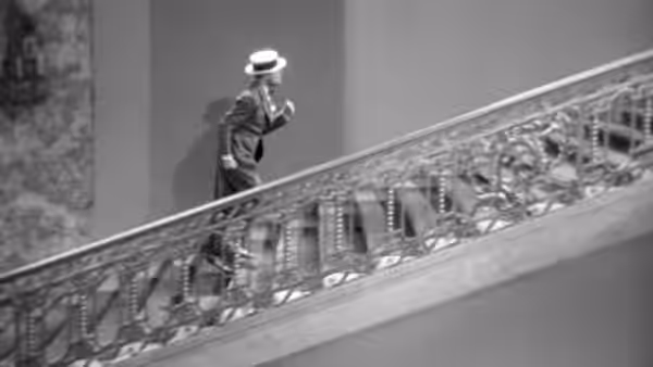 Maurice Courtelin in a hat stands above a shopfront, gesturing from an elevated walkway