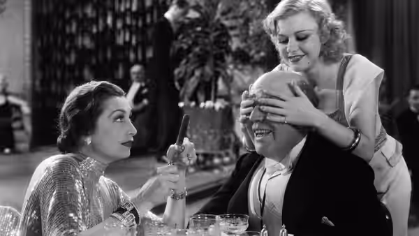 A woman covers a seated man's eyes while another watches from the table