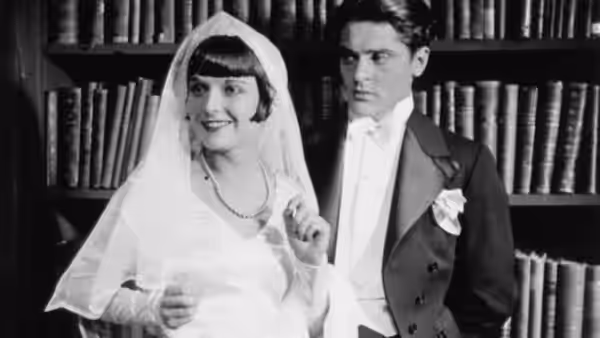 Lulu (Louise Brooks), dressed as a bride, stands before bookshelves beside Alwa Schön (Francis Lederer)