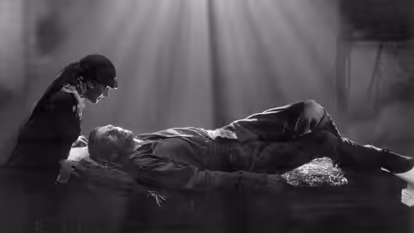 A man leans over another man lying on a narrow bed beneath a shaft of light