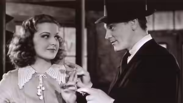 A suited man in a fedora reaches toward a woman’s collar indoors
