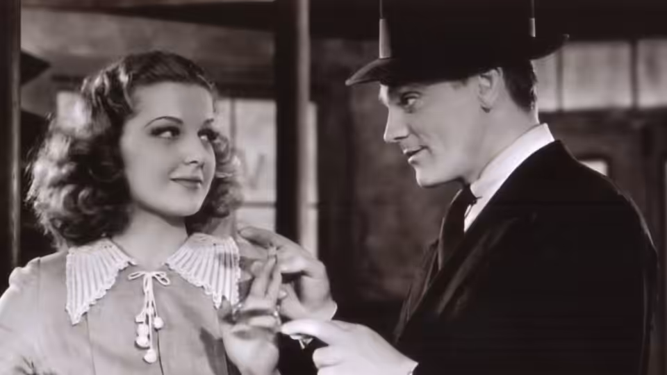 A suited man in a fedora reaches toward a woman’s collar indoors