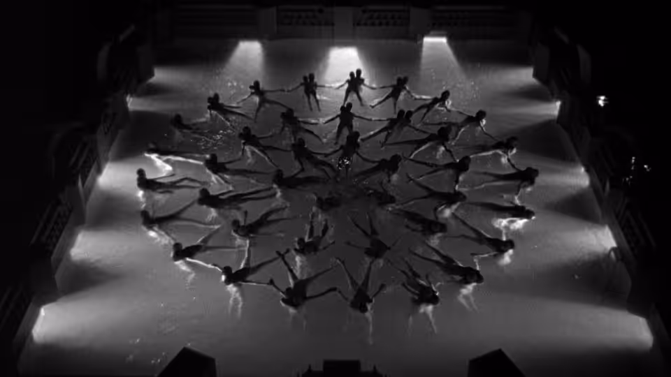 A chorus line forms a geometric pattern across the stage under spotlights