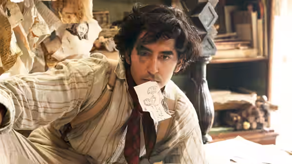 David Copperfield leans over a desk with a paper held between his lips