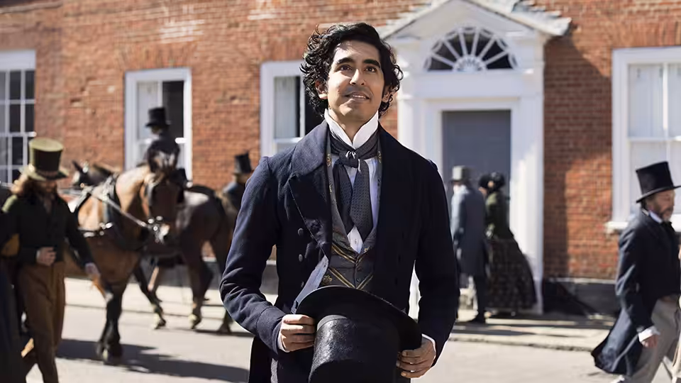 David Copperfield (Dev Patel) stands outside a brick house, holding his top hat as people and a horse pass behind him