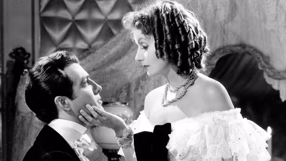 Marguerite Gautier (Greta Garbo) cups Armand Duval’s face in an elegant room