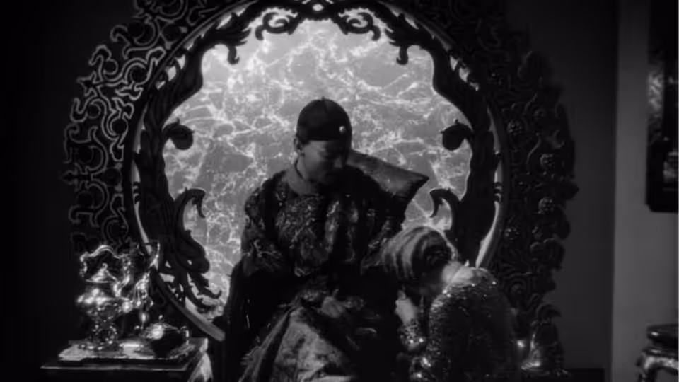 A man in an embroidered robe sits behind an ornate screen as a woman faces him