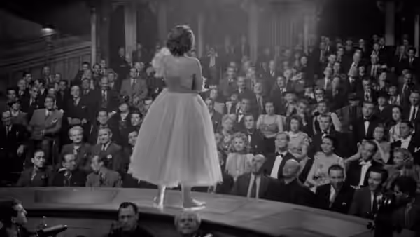 A ballerina faces rows of seated men from the edge of a stage