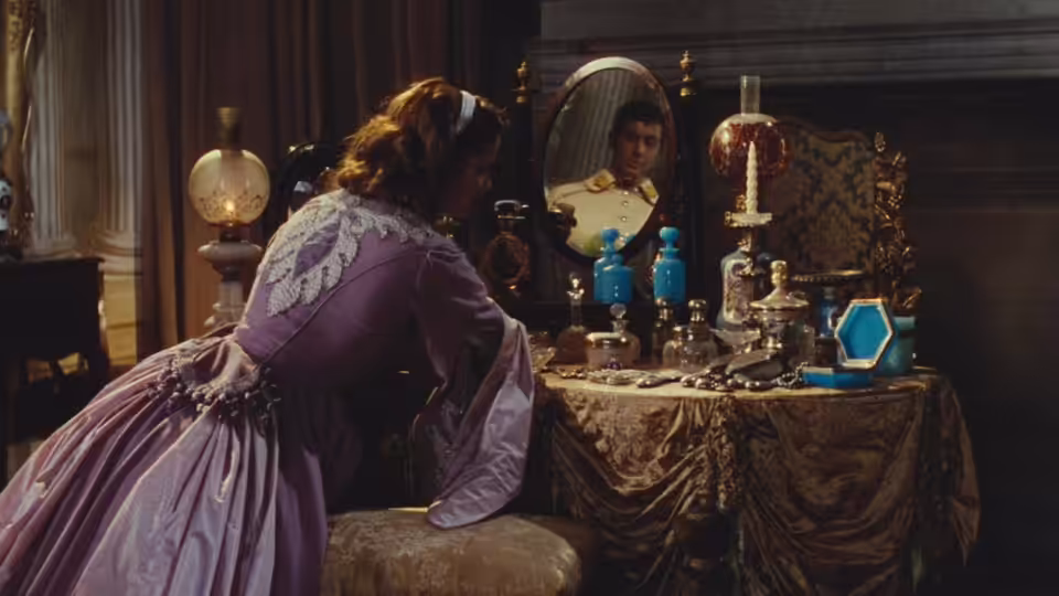 A woman in a lavender gown sits at a dressing table, studying her reflection
