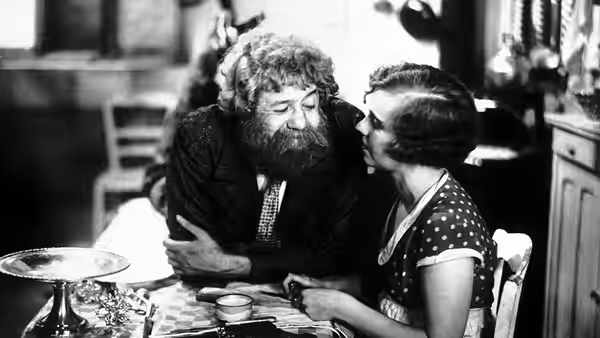 Boudu (Michel Simon) sits at a table as a woman leans toward him