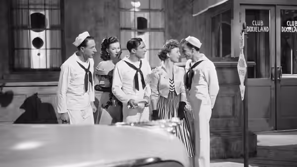 Three sailors talk with a woman in a striped skirt outside a city storefront