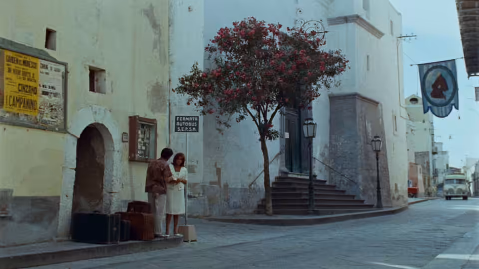 Two women stand beside luggage on a quiet street lined with white buildings