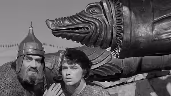 An older armored man stands beside a younger man beneath an enormous cannon
