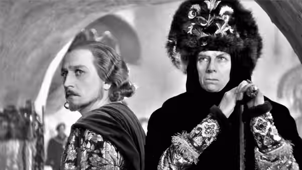 Two elaborately dressed men stand together, one wearing a towering fur headdress