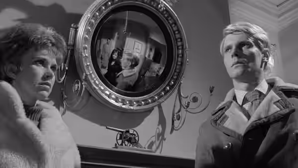 A woman and a man stand beside a large circular mirror reflecting figures inside a room