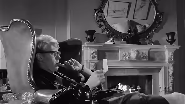 A man wearing glasses smokes in an armchair and holds a small paper by the fireplace