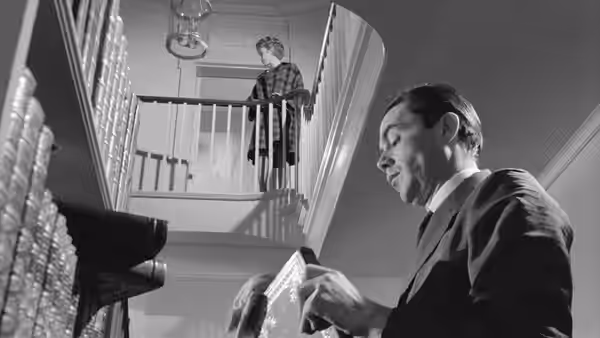 A man reads a paper below another man standing on an upstairs landing