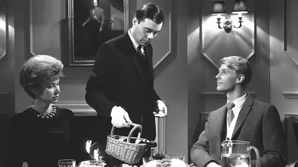 Hugo Barrett (Dirk Bogarde) serves at the table while Tony and Susan look on