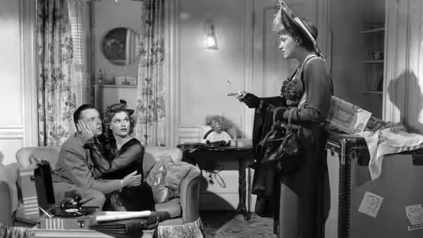 A seated couple watch a woman in a hat standing beside their living room sofa