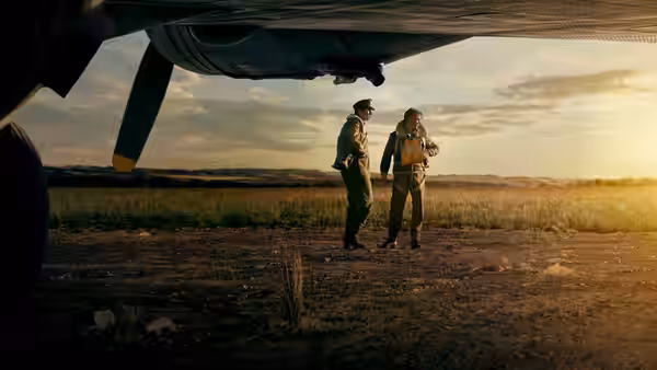 Two airmen stand beneath a bomber wing on a sunlit airfield