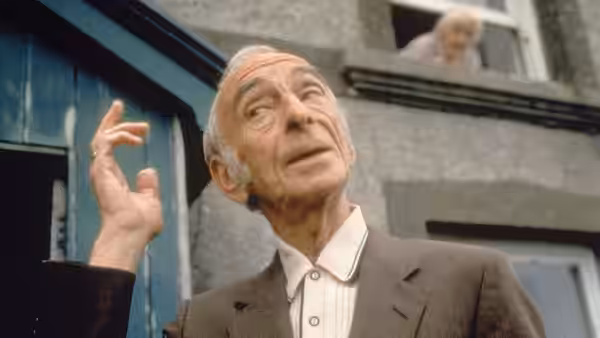 Michael O'Sullivan (David Kelly) gestures outside a house as someone watches from an upstairs window