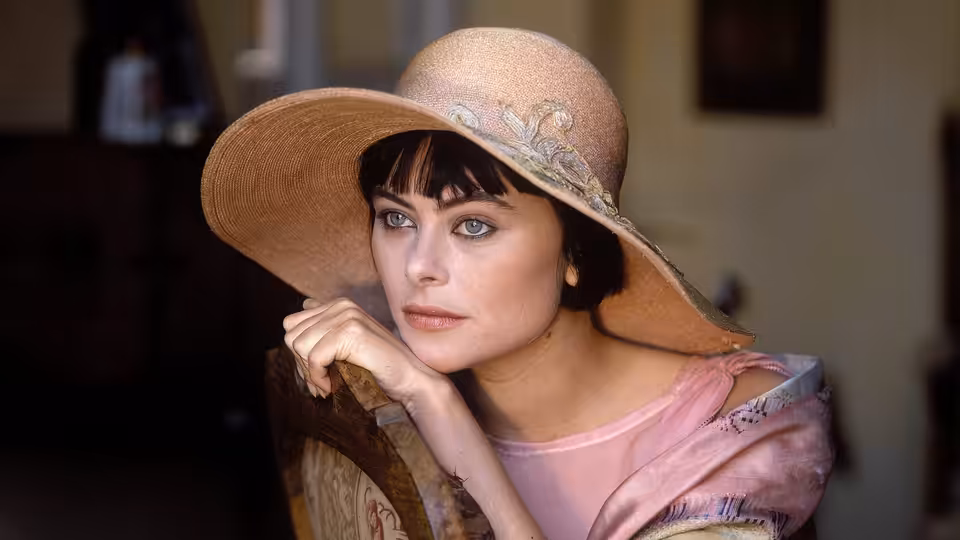 Caroline Dester (Polly Walker) leans on a chair in a wide-brimmed hat