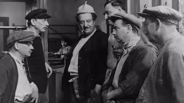 Villagers gather around a man in a dark jacket inside a room