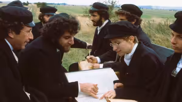 Yentl studies an open book with several young men gathered around a table outdoors