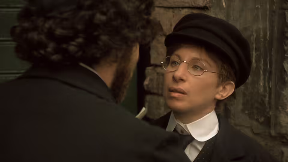 Yentl, dressed as a young man, speaks with a bearded man beside a doorway