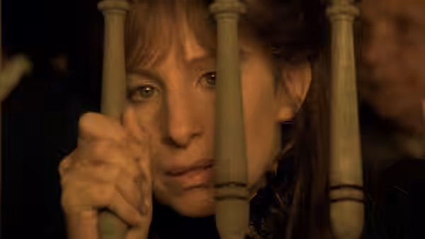A woman peers through wooden bars, gripping one as another figure stands behind her