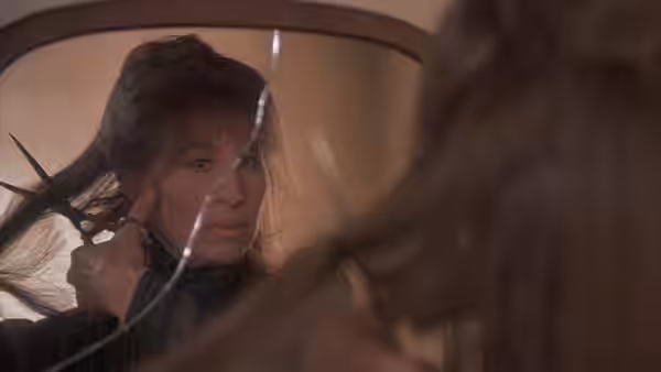 A woman looks into a mirror as someone holds scissors beside her hair