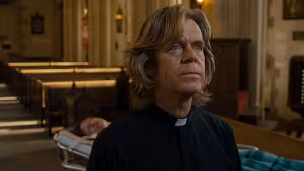 Father Brendan (William H. Macy) gazes upward in church as Mark (John Hawkes) lies on a stretcher