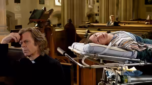 Father Brendan (William H. Macy) sits beside Mark (John Hawkes), who lies on a stretcher in church