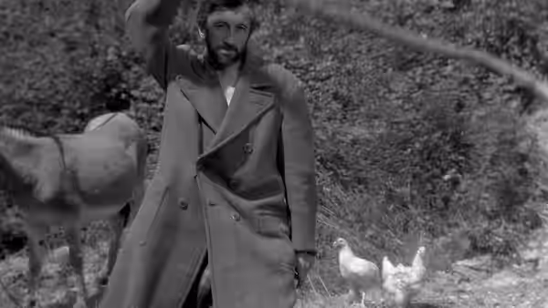A man stands beside a donkey and raises one arm as chickens gather nearby