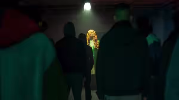 Several people watch a brightly dressed figure from a dim corridor