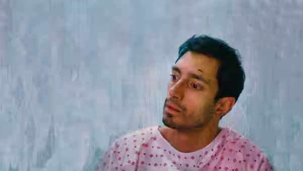 Zed (Riz Ahmed) in a patterned hospital gown looks to the side