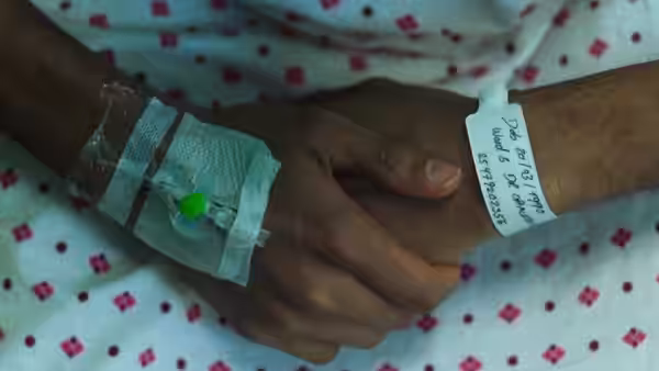 A patient’s hands rest together, one bearing an IV and hospital wristband