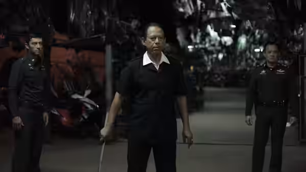 Chang (Vithaya Pansringarm) stands holding a sword between two uniformed men in a garage