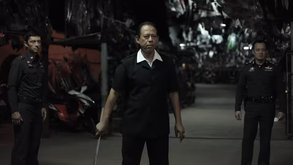 Chang (Vithaya Pansringarm) stands holding a sword between two uniformed men in a garage