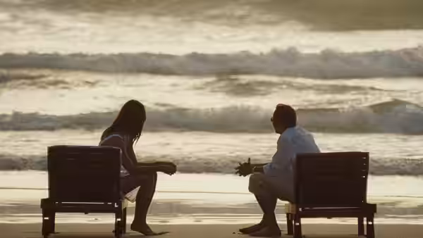 A man and woman sit facing each other beside the ocean