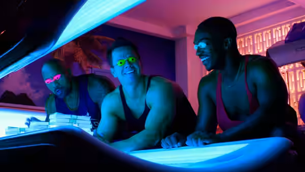 Three muscular men lie beneath ultraviolet lights in a tanning salon