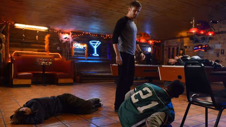 David (Dan Stevens) stands over two fallen men in a neon-lit bar