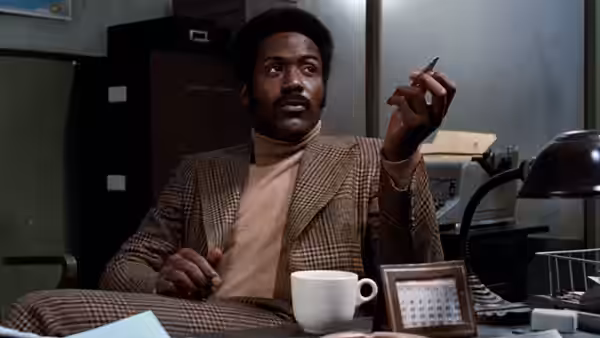 John Shaft (Richard Roundtree) sits at a desk, holding a cigarette beside a coffee cup